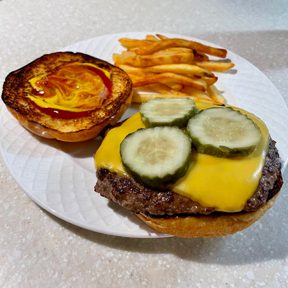 Grass fed hamburger patty with cheese and pickles on a bun, served with French fries on a white plate.