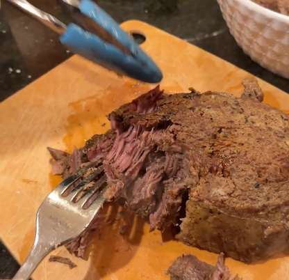 Close-up of grass fed arm roast being cut with tongs and a fork on a wooden cutting board.